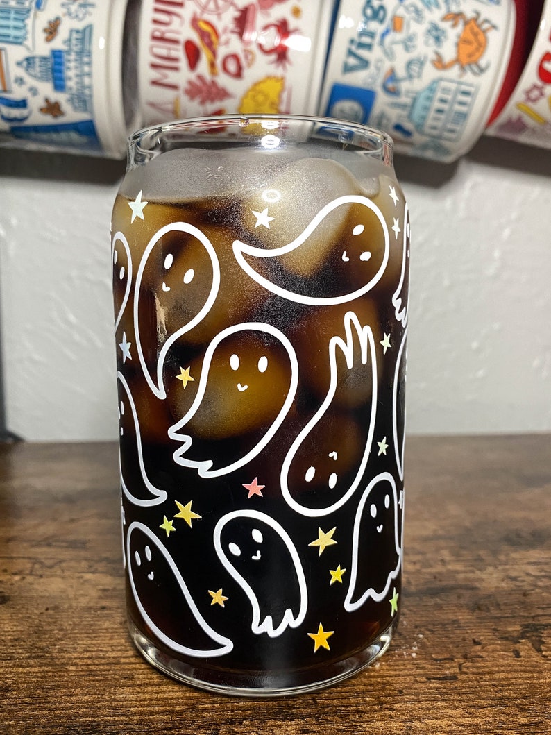 May include: A glass with a black and white ghost pattern and a dark brown liquid inside. The glass has a white rim and a clear base. The ghosts are outlined in white and have small eyes and smiles. There are also small white stars scattered around the ghosts.