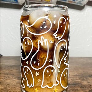 May include: A glass with a white ghost pattern and gold stars filled with iced coffee. The glass is on a wooden surface.