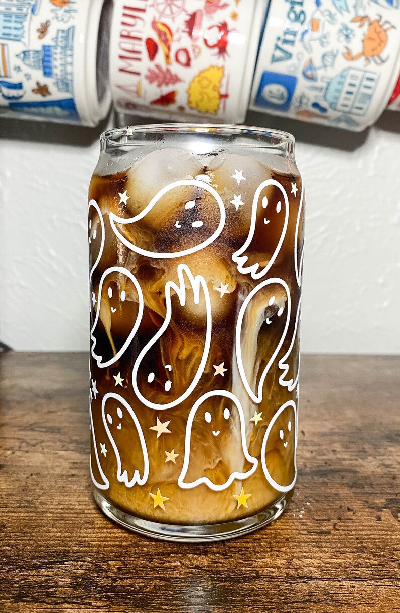 May include: A glass with a white ghost pattern and gold stars filled with iced coffee. The glass is on a wooden surface.