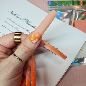 Peut inclure: Des ongles press-on orange et rose avec des paillettes et des motifs floraux. Les ongles sont longs et de forme carr&eacute;e. Le texte "Nails by xX Honeebec" est &eacute;crit sur une surface blanche.