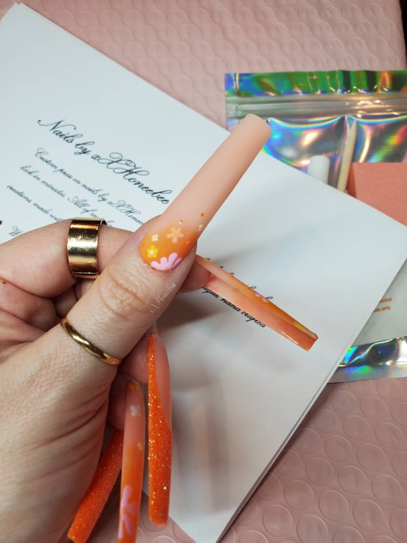 Peut inclure: Des ongles press-on orange et rose avec des paillettes et des motifs floraux. Les ongles sont longs et de forme carr&eacute;e. Le texte "Nails by xX Honeebec" est &eacute;crit sur une surface blanche.