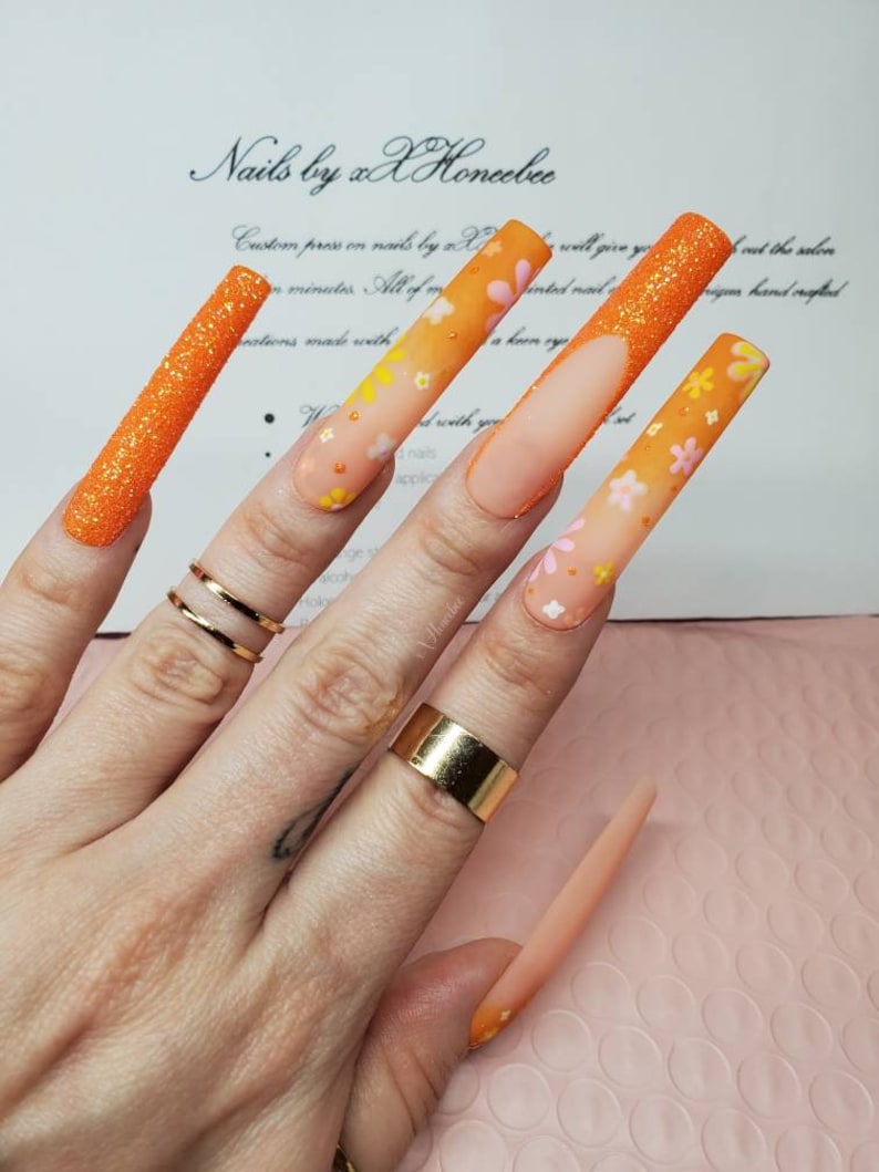 Peut inclure: Longues ongles en gel avec un design floral, de couleur orange et blanche. Les ongles sont ombr&eacute;s avec une pointe paillet&eacute;e. Le texte "Nails by xX Honcebee" est visible en arri&egrave;re-plan.
