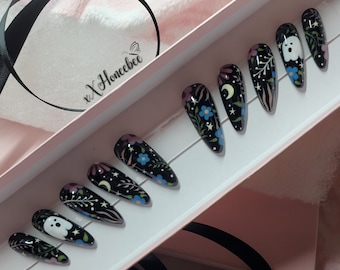 May include: A set of ten black press-on nails with a spooky Halloween design. The nails feature a black base with white ghosts, blue flowers, green leaves, and gold stars. The nails are in a white box with the text "xx Honeebee" on the lid.