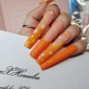 Peut inclure: Ongles longs en amande avec une finition scintillante orange et blanche et un motif floral.