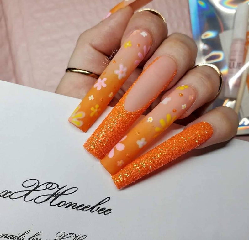Peut inclure: Ongles longs en amande avec une finition scintillante orange et blanche et un motif floral.