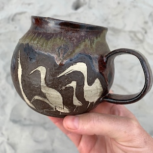 Puede incluir: Una taza de cerámica marrón con un esmalte blanco y un diseño de pájaros en vuelo. La taza tiene un cuerpo redondeado y un asa.