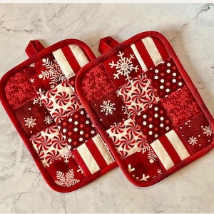 May include: Two red and white quilted pot holders with a Christmas theme. Each pot holder features a patchwork design with peppermint swirls, snowflakes, and polka dots. They have red binding and a loop for hanging.
