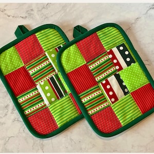 May include: Two square quilted potholders with a patchwork design. The potholders feature red, green, and white fabrics in polka dot, striped, and solid patterns. Each potholder has a green border and a green loop for hanging.
