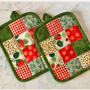 May include: Two square quilted pot holders with a green border and hanging loop. The patchwork design features red gingham, strawberries, floral patterns, and green foliage. These kitchen accessories are designed to protect hands from heat.