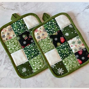 May include: Two square quilted potholders with a green border and hanging loop. The patchwork design features white, green, and black squares with snowflake and mitten patterns. The potholders are designed for kitchen use.
