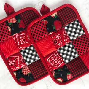 May include: Two red quilted pot holders with a patchwork design featuring black and white gingham, red polka dots, red bandana print, and black fabric with red apples and letters.