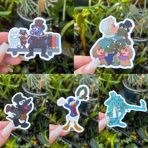 May include: A collection of colorful stickers featuring various cartoon characters. The stickers include Gonzo reading a book, a group of Muppets, a crow in a coat, Donald Duck with a wreath, and a blue ghost-like figure.