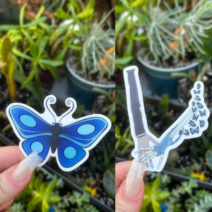 May include: Two vinyl stickers, one featuring a blue butterfly with white accents and the other a skeletal hand reaching out with a trail of blue butterflies behind it.
