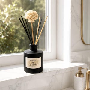 Può includere: Un flacone diffusore a bastoncini in vetro nero opaco con un'etichetta color crema che riporta la scritta Vanilla & Lavande. Il flacone contiene diversi bastoncini neri e beige, uno dei quali sormontato da un fiore decorativo color crema.