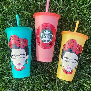 May include: Three reusable cups with lids and straws. The cups are pink, turquoise, and yellow. Each cup has a black and white illustration of Frida Kahlo with flowers in her hair. The pink cup has a red circle with the text "Fragile Like a Bomb" in white.