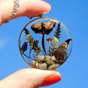 May include: A clear, round pendant necklace with a silver chain. The pendant contains a miniature scene with a mushroom, small plants, and a snail shell, all encased in clear resin. The background is a bright blue sky.