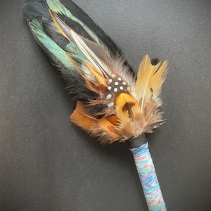 May include: A ceremonial fan featuring a cluster of layered feathers in shades of black, iridescent green, orange, and brown, with white-spotted accents. The handle is wrapped in a multi-coloured, textured yarn in hues of blue, pink, and white.
