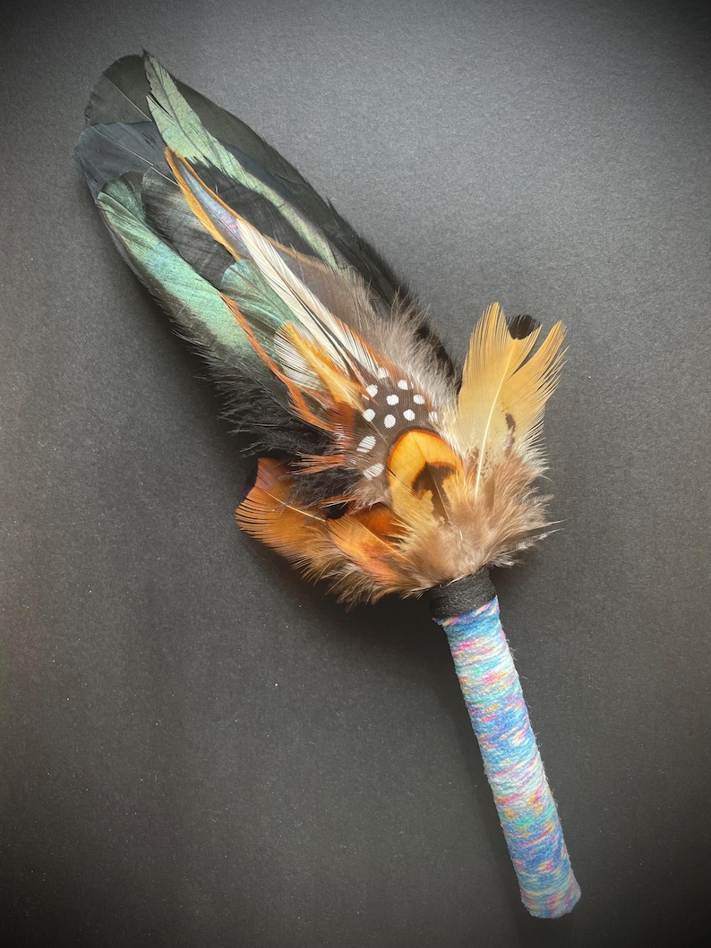 May include: A ceremonial fan featuring a cluster of layered feathers in shades of black, iridescent green, orange, and brown, with white-spotted accents. The handle is wrapped in a multi-coloured, textured yarn in hues of blue, pink, and white.