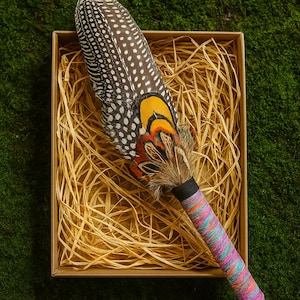 May include: A ceremonial rattle featuring a large, spotted brown and white guinea fowl feather, accented with vibrant yellow, orange, and black pheasant feathers. The handle is wrapped in a multi-coloured, textured thread in shades of pink, blue, and orange.