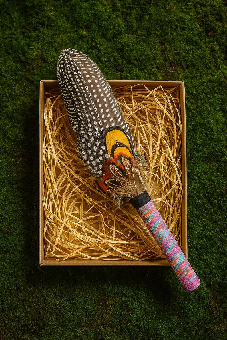 May include: A ceremonial rattle featuring a large, spotted brown and white guinea fowl feather, accented with vibrant yellow, orange, and black pheasant feathers. The handle is wrapped in a multi-coloured, textured thread in shades of pink, blue, and orange.