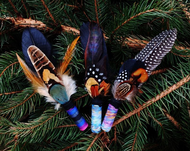 May include: Three decorative feather bundles featuring a mix of dark, iridescent, and spotted plumage. Each bundle is secured with a tightly wrapped handle in vibrant blue, purple, and multi-coloured thread, creating a distinct textured base.