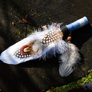 May include: A ceremonial fan featuring a handle wrapped in blue and white fabric, adorned with a black band and wooden beads. The fan is decorated with a large white feather, spotted guinea fowl feathers, and a pheasant feather with red accents.