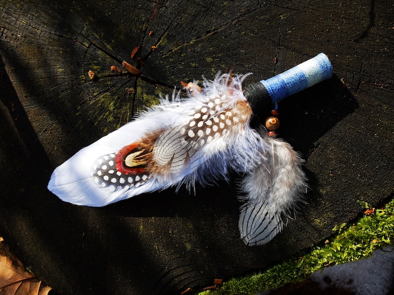 May include: A ceremonial fan featuring a handle wrapped in blue and white fabric, adorned with a black band and wooden beads. The fan is decorated with a large white feather, spotted guinea fowl feathers, and a pheasant feather with red accents.