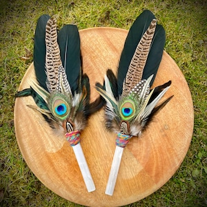 May include: Two decorative feather wands with peacock and other feathers, featuring a vibrant blue eye. The wands have white handles wrapped with colourful thread. They are displayed on a round wooden tray, set against a backdrop of green grass.