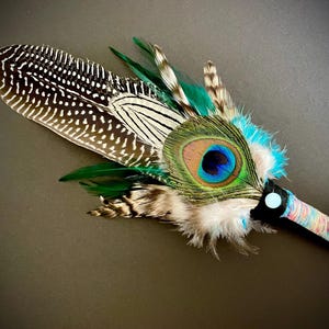 Könnte beinhalten: Ein dekorativer Zauberstab mit einem Pfauenfedernauge, akzentuiert mit grünen, blauen und braunen Federn. Der Griff ist mit farbigem Garn umwickelt. Der Zauberstab ist vor einem dunklen Hintergrund platziert.