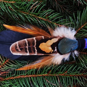May include: A decorative fan featuring a layered arrangement of black, brown, and tan feathers with a handle wrapped in black, blue, and magenta thread. The feathers display distinct striped and solid patterns with soft downy accents.