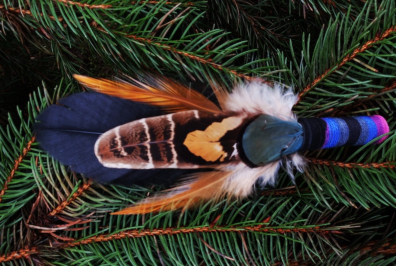 May include: A decorative fan featuring a layered arrangement of black, brown, and tan feathers with a handle wrapped in black, blue, and magenta thread. The feathers display distinct striped and solid patterns with soft downy accents.