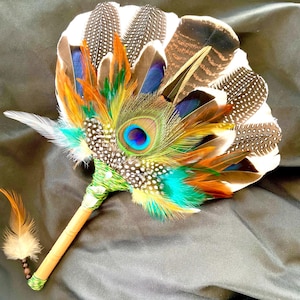 May include: A handheld fan constructed from a variety of feathers, including a central peacock eye feather, spotted guinea fowl feathers, and orange and turquoise accents. The handle is wrapped in green thread with a small dangling feather.