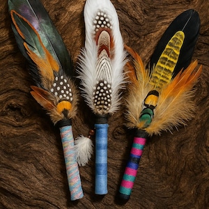 May include: Three decorative feather fans featuring long, layered plumes in shades of white, black, orange, and iridescent green. Each handle is wrapped in colourful thread, with patterns including solid blue, multi-coloured stripes, and pink and teal bands.