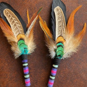 May include: Two feather fans featuring long black, brown, and orange feathers layered with iridescent green and teal plumage. The handles are wrapped in black, purple, pink, and light green yarn, creating a striped, colourful pattern.