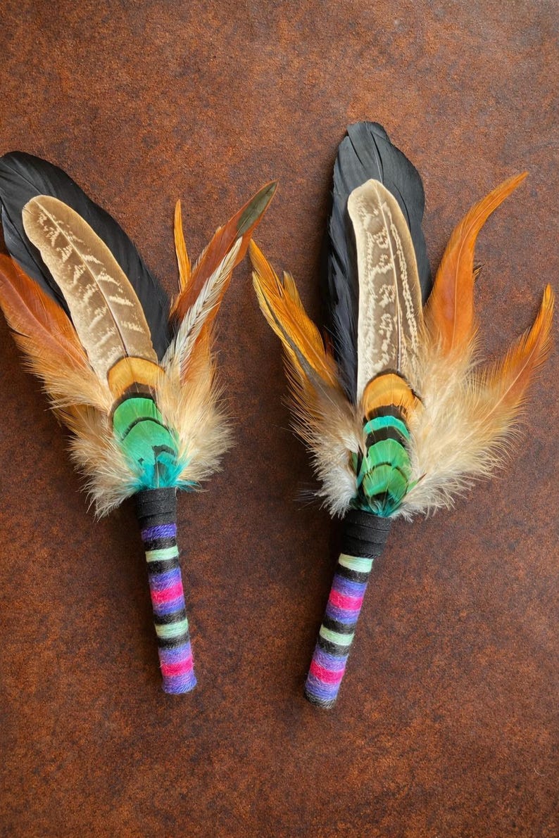 May include: Two feather fans featuring long black, brown, and orange feathers layered with iridescent green and teal plumage. The handles are wrapped in black, purple, pink, and light green yarn, creating a striped, colourful pattern.