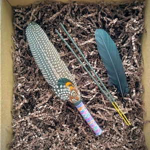 May include: A decorative fan made from a large, spotted guinea fowl feather with orange and green accents at the base, featuring a handle wrapped in multicoloured thread, accompanied by a single dark feather and two thin green sticks with yellow tips.