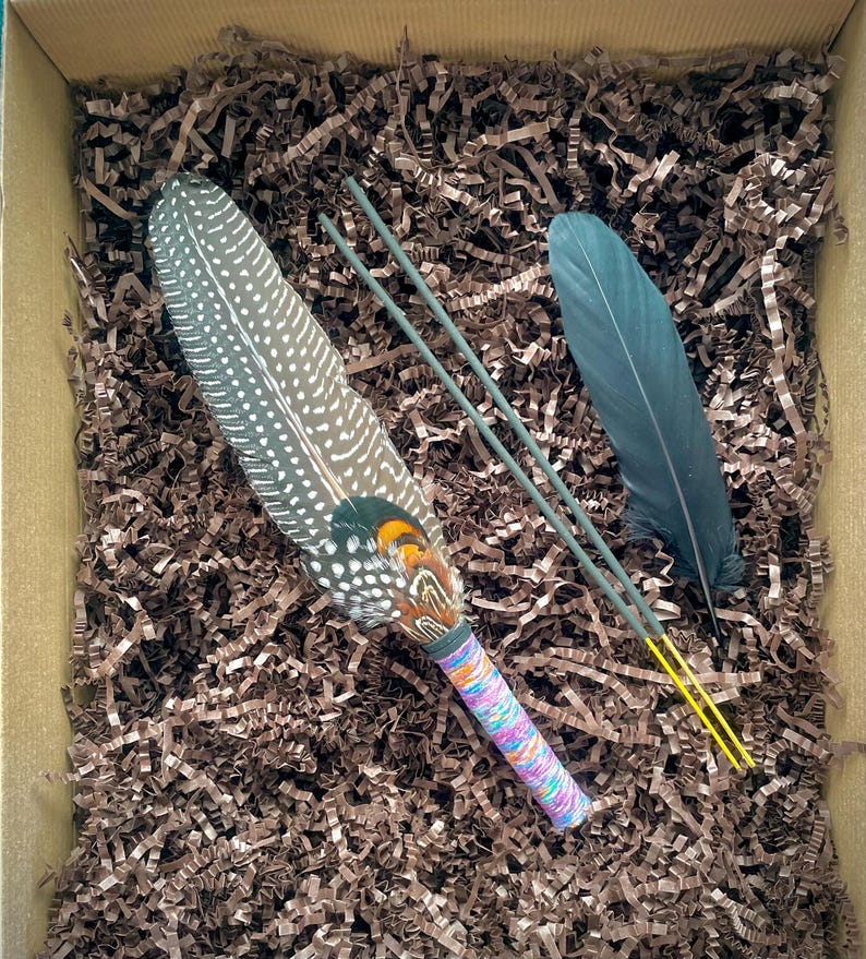 May include: A decorative fan made from a large, spotted guinea fowl feather with orange and green accents at the base, featuring a handle wrapped in multicoloured thread, accompanied by a single dark feather and two thin green sticks with yellow tips.