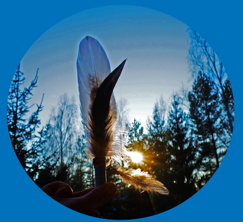 May include: A feather ornament featuring a central white and brown plume attached to a cylindrical base, with a smaller secondary feather and a metallic bead accent dangling from the side, backlit by a bright sun in a forest setting.