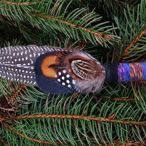 May include: A decorative feather wand featuring a long, spotted guinea fowl feather layered with brown, black, and orange pheasant feathers. The handle is wrapped in a vibrant, multi-coloured purple, blue, and orange yarn with a black band.
