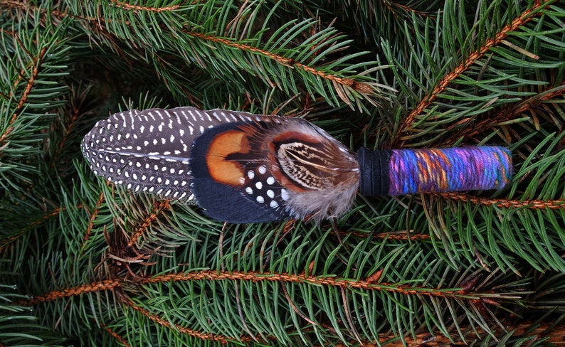 May include: A decorative feather wand featuring a long, spotted guinea fowl feather layered with brown, black, and orange pheasant feathers. The handle is wrapped in a vibrant, multi-coloured purple, blue, and orange yarn with a black band.