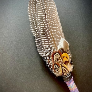 May include: A decorative fan made of brown and white spotted guinea fowl feathers, accented with smaller orange, black, and white pheasant feathers at the base. The handle is wrapped in a textured, multi-coloured purple and blue yarn.