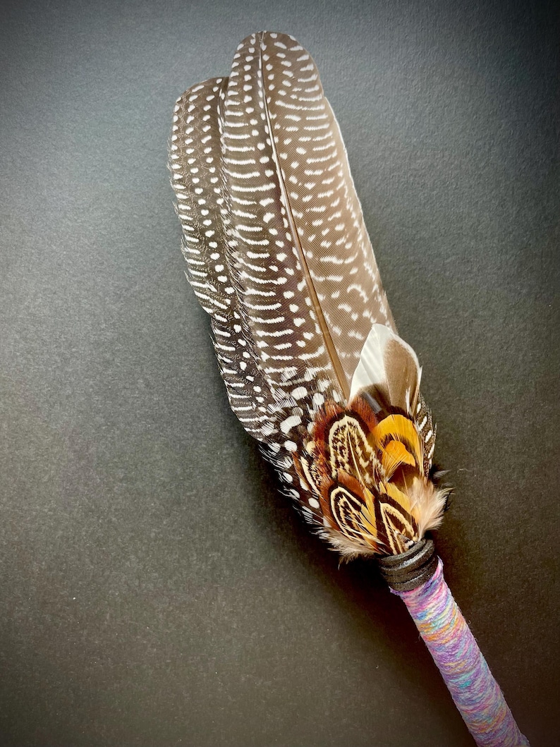 May include: A decorative fan made of brown and white spotted guinea fowl feathers, accented with smaller orange, black, and white pheasant feathers at the base. The handle is wrapped in a textured, multi-coloured purple and blue yarn.