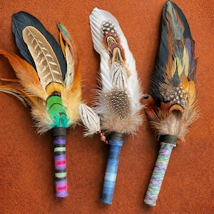 May include: Three handcrafted feather fans featuring layered plumage in shades of black, white, brown, and iridescent green. Each fan has a handle wrapped in colourful, multi-textured yarn, showcasing distinct striped and variegated patterns.
