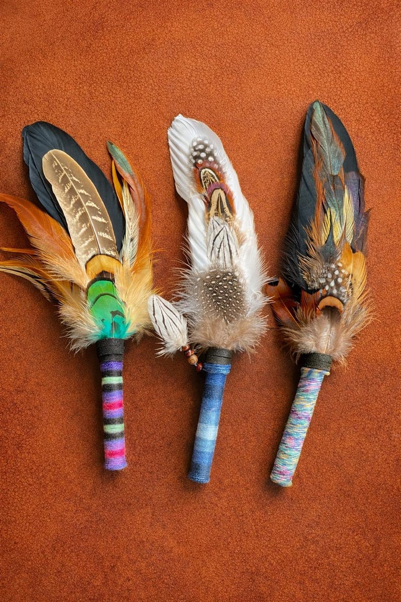 May include: Three handcrafted feather fans featuring layered plumage in shades of black, white, brown, and iridescent green. Each fan has a handle wrapped in colourful, multi-textured yarn, showcasing distinct striped and variegated patterns.