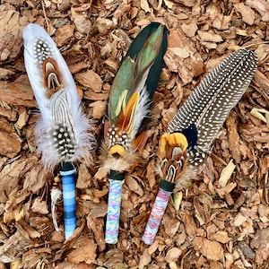 May include: Three decorative feather wands featuring layered plumage in white, brown, black, and iridescent green. Each wand has a wrapped handle in blue stripes, multicoloured speckles, or pink and white patterns with small beads.