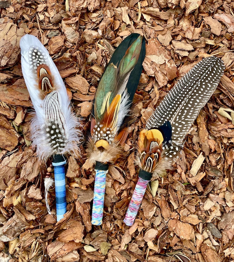 May include: Three decorative feather wands featuring layered plumage in white, brown, black, and iridescent green. Each wand has a wrapped handle in blue stripes, multicoloured speckles, or pink and white patterns with small beads.