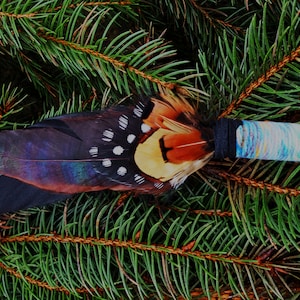 May include: A decorative wand featuring a cluster of dark brown, iridescent black, and spotted pheasant feathers attached to a handle wrapped in black thread and a light blue, white, and yellow variegated yarn.