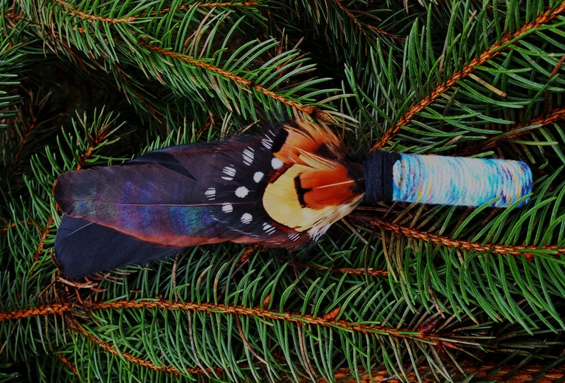 May include: A decorative wand featuring a cluster of dark brown, iridescent black, and spotted pheasant feathers attached to a handle wrapped in black thread and a light blue, white, and yellow variegated yarn.