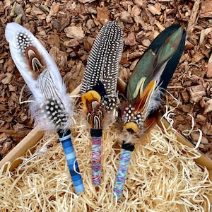 May include: Three decorative feather wands featuring layered plumage in white, brown, black, and iridescent green. Each handle is wrapped in colourful thread, with the left wand featuring blue stripes and the others displaying multicoloured patterns.