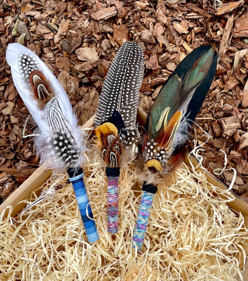May include: Three decorative feather wands featuring layered plumage in white, brown, black, and iridescent green. Each handle is wrapped in colourful thread, with the left wand featuring blue stripes and the others displaying multicoloured patterns.